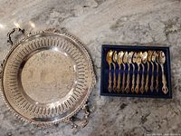 Full view of the silver plated round serving tray with ornate handles and a set of 10 gold-tone spoons in velvet-lined box
