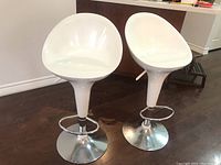 Full view of two white bar stools with molded plastic seats and chrome bases positioned on a hardwood floor.