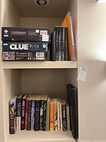 Shelf with stacked Game of Thrones Monopoly, Clue board games, Eurographics puzzle box, and black CD cases, alongside an orange book.