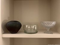 Three items on a shelf: a dark matte vase, a clear glass bowl, and a Czech crystal footed bowl with cut patterns on a square base.