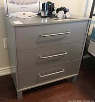 Full front view of gray laminate end table with three metal handle drawers and items placed on top.