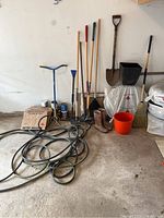 Wide view showing garden tools including rakes, shovels, broom, rain boots, planter, bucket, hose on garage floor.
