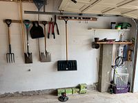 Garage wall with hanging garden tools including pitchfork, shovels, dustpan, and garden shears. Boxes of garden supplies on the floor and various containers on metal shelves.