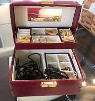 Closed red leather jewelry box showing its mirror and interior compartments with several pieces of jewelry inside.