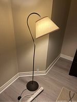 Full view of black metal floor lamp with beige fabric lampshade and power cord on floor showing overall condition and style