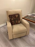 Front and angle view of beige Lazboy reclining armchair with floral pillow on light grey wood floor near tan wall.