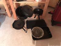 Photo of assorted black pots, pans, and bakeware arranged on floor including enamel roasting pan with lid, frying pans, cast iron pan, oven top griddle, bunt pan and open roast pan.