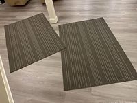 Two matching brown area rugs with vertical stripe pattern laid on light wood floor, one slightly folded at a corner