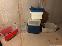 Overview of multiple plastic tote bins stacked and arranged against wall in basement, showing various sizes and colors.
