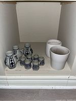 Six ceramic West German shot glasses with blue diamond pattern, three small matching pitchers, and two white ceramic planters arranged on a shelf.