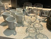Five white ceramic mugs and five clear stemware wine glasses on a counter.