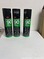 Three new unopened cans of 3M Hi-Strength 90 Spray Adhesive with green and black colored labels, standing upright on a white surface.