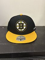 Front view of the Boston Bruins hat, showing the logo and sticker on the yellow brim.