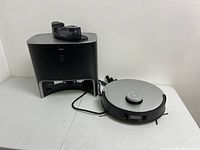 Robot vacuum and charging station side by side on white table, cables visible