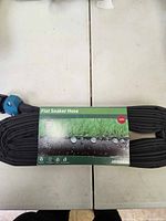 Photo of flat soaker hose coiled with label showing product name, water droplets on grass graphic, and length of 25 feet.
