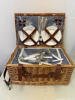 Open picnic basket showing contents: two white plates secured with leather straps on lid and multiple wrapped utensils inside basket compartment.
