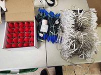 Box with string of large red Christmas bulb lights, blue C9 bulb string, and several bundles of white mini Christmas lights on table.