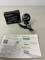 Mini WiFi camera with USB cable, instruction manual, and box on a white surface.