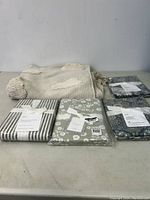 Four folded Pottery Barn king size shams with tags and cream H&M rug with textured designs and fringe edges on a table.