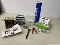 Photo showing all items in the lot arranged on a tabletop including grease tube, pliers, hammer, metal part, boxes, syringe and instruction sheet.