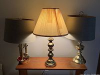Three table lamps on a wooden surface; middle lamp lit with beige pleated shade, two side lamps with gray shades and brass bases.