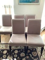 Front and angled view of five beige upholstered dining chairs with dark wooden legs, arranged on a decorative rug with visible stains and discoloration on fabric.
