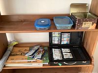 View of a shelf with two black cases of paints, plastic containers with unknown contents, several sketchbooks, and loose papers