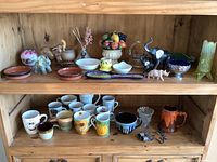Wide shot of three shelves containing various small decor items, animal figurines, mugs, bowls, and dishes packed densely showing variety and condition, some items with visible chips.