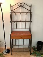 Front view of the bakers rack showing iron frame with scroll design, wooden drawer with visible surface wear, and wire shelves.