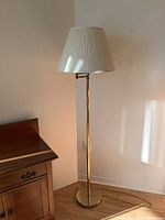 Full view of the floor lamp showing brass stem and base, white pleated shade, placed beside wooden furniture.