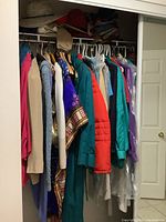 Wide view of closet with multiple vintage ladies clothing items hung including coats, jackets, sweaters, dresses, blouses, scarves, and visible hats on the shelf above.