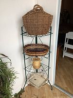 Full view of metal corner plant stand with large woven basket on top shelf, oval basket in middle, and metal tray basket at bottom. Placed near a corner with light wooden floor.