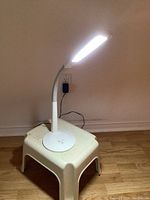 Full view of the white OttLite desk lamp on a small step stool against a wall, showing the lamp switched on.