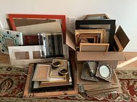 Wide view of a large collection of picture frames stacked on floor and inside a cardboard box, showing various colors, sizes, and styles.