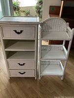 Pair of white bathroom storage shelves placed indoors; left unit is taller with drawers and shelves, right unit is shorter with wicker style shelves and backrest detail.