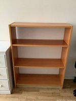 Front view of the small wooden bookcase showing three shelves and no backing, with wood finish and situated beside white drawer unit and chair.