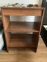Front view of small storage shelf showing three shelves and wood laminate finish with wear marks.