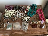 Assorted vintage costume jewelry including multiple necklaces, bracelets, earrings, and two pairs of eyeglasses displayed on a wooden surface.