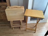 Four folding wooden TV tables stacked in a foldable wooden stand with one additional folding table beside it against a wood floor background.