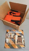 Open brown cardboard box containing neatly folded bright orange moving straps with black edges. Instruction manual placed next to the box.