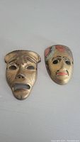 Front view of two brass masks, one with distressed expression, the other painted with red and black details