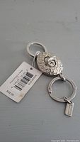 Photo of metallic silver Coach keychain, showing front side with oval textured plate and attached price tag.