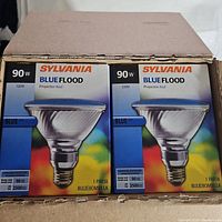 Partial top view of the open shipping case showing six boxes of Sylvania 90W 120V Blue Flood light bulbs inside, boxes are sealed and new.