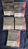 Five sealed packages of Zebra Doodler'z gel stick pens, each pack holding 10 pens in multiple colors including black, blue, red, and purple ink, arranged on a dark cloth.