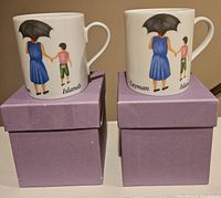 Pair of white porcelain mugs with painted designs of a woman and child holding hands under umbrella, labeled Cayman Islands, each on top of lavender box.