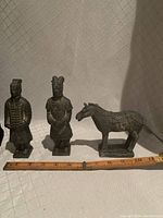 Photo of two terracotta warrior figurines and one horse figurine beside a wooden ruler showing measurements.