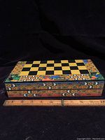 Closed colorful decorated wooden chessboard box with Peruvian style motifs shown next to a ruler for size reference, about 10x10 inches.
