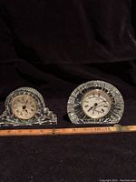 Two crystal clocks side by side on dark cloth background with wooden ruler showing size.