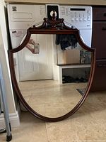 Frontal photo of the vintage mahogany framed crest mirror against a tiled floor showing the top carved crest and shield-shaped reflective glass.
