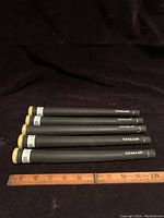 Photo showing five black golf iron grips with yellow end caps placed next to a ruler indicating approximately 10 inches length